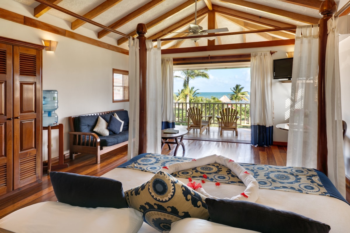 A comfortable bedroom features a spacious bed adorned with decorative pillows. A seating area with a couch is visible, alongside a balcony with wooden chairs offering views of the Caribbean Sea. Light streams in through sheer curtains, enhancing the serene atmosphere.