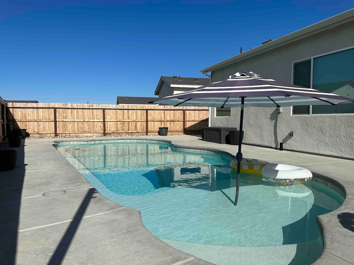 A private pool area is shown, featuring a clear blue pool surrounded by a concrete patio. An umbrella is positioned over a lounge chair, offering shade. The backyard is enclosed by a wooden fence, providing a secluded feel.