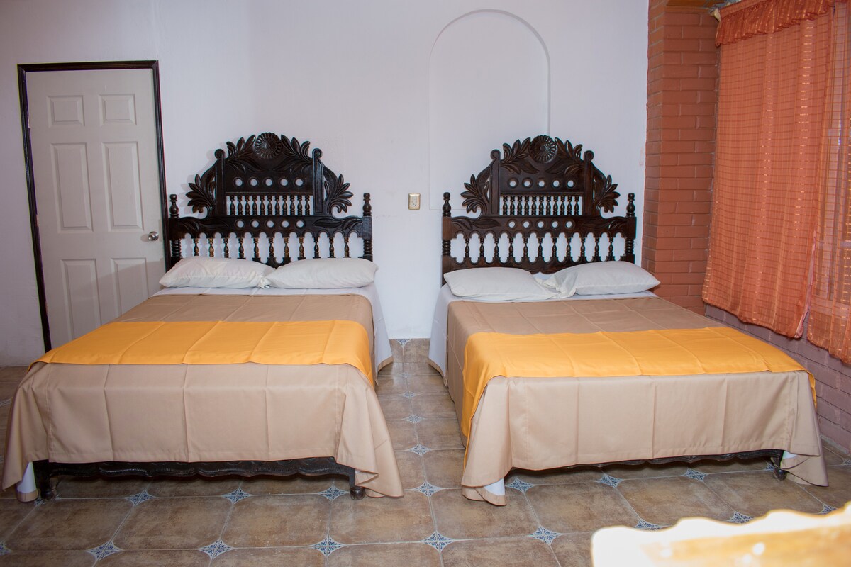 A spacious room is furnished with two large beds featuring ornate wooden headboards. Each bed is dressed with simple beige and orange bedding. Natural light filters through orange curtains, and a door is visible in the background, contributing to the room's welcoming layout.