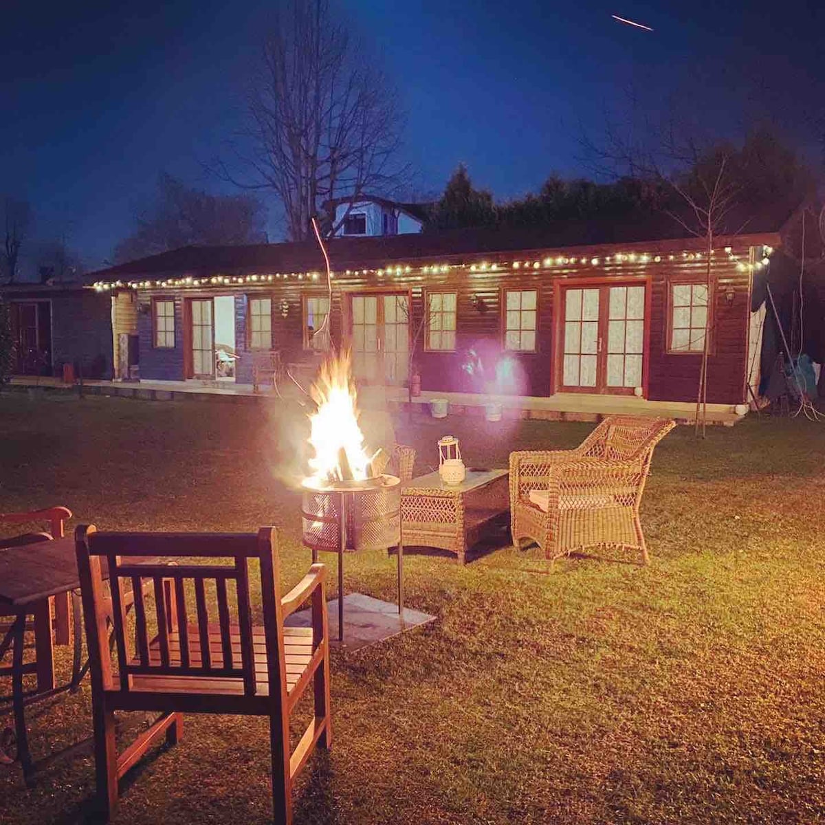 The outdoor area features comfortable rattan chairs arranged around a fire pit, creating a cozy setting. Soft lights outline the cabin, providing illumination against the evening sky. The spacious lawn is well-maintained, inviting relaxation and gatherings.