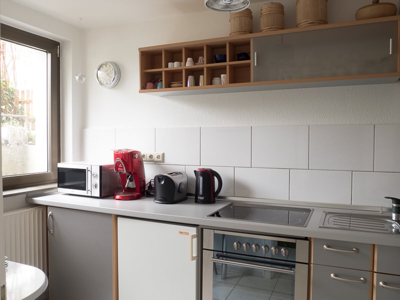 The kitchen features a combination of modern appliances, including a microwave, coffee maker, and kettle. A contemporary stovetop and oven are also present, alongside a neatly organized shelf displaying cups. A window allows natural light to brighten the space.