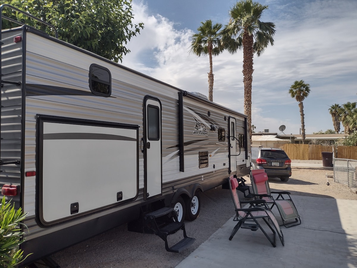 An RV is positioned under palm trees, with a shaded area featuring two lounge chairs. A vehicle is parked nearby, and the background reveals a fenced area, indicating a spacious outdoor setting.