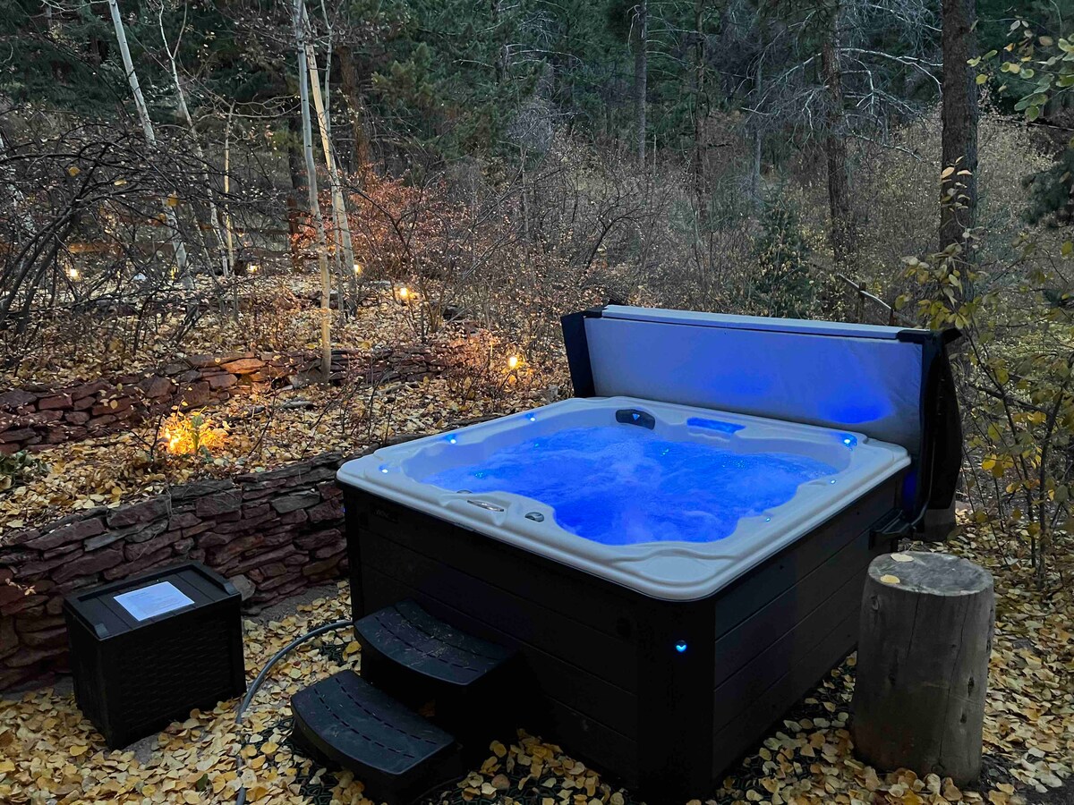 A private hot tub is nestled among golden autumn leaves, illuminated by soft blue lights. The surrounding area features natural foliage and scattered lights that enhance the tranquil setting, creating a serene oasis for relaxation.