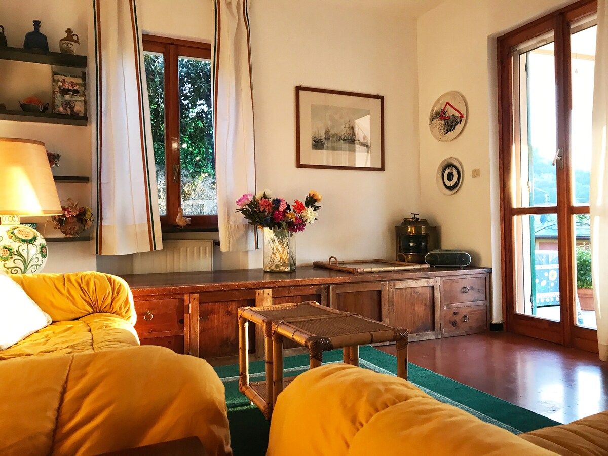 A cozy living area is presented with warm yellow sofas and a wooden coffee table. Natural light enters through large glass doors, creating a bright environment. Shelves with decorative items are visible, along with a flower arrangement that adds a touch of color.