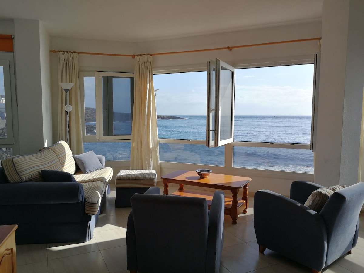 The living area features large windows providing expansive views of the ocean. Comfortable seating arrangements include two armchairs and a sofa. A wooden coffee table is situated in the center, while sheer curtains allow natural light to fill the space.