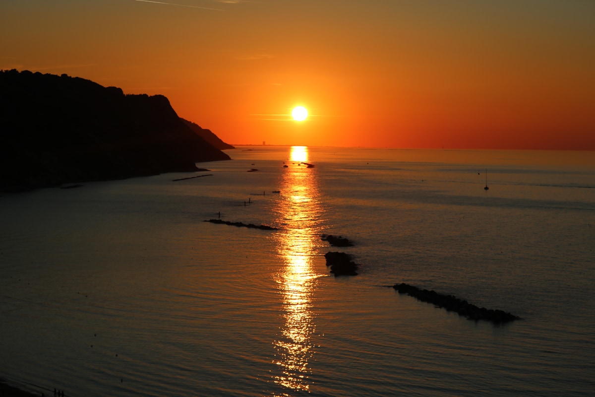 A serene sunset is captured over a calm sea, reflecting warm hues of orange and gold across the water. Silhouettes of gentle cliffs frame the horizon, while small boats can be seen in the distance, contributing to the tranquil coastal scene.
