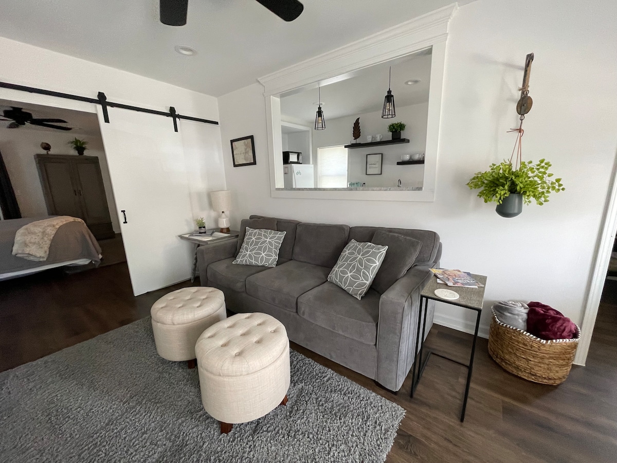 A cozy living area features a gray sofa adorned with two decorative pillows. Two upholstered ottomans provide additional seating. A small side table holds a lamp and decorative items. A sliding door and a mirror add depth to the room, which is anchored by a soft gray rug.
