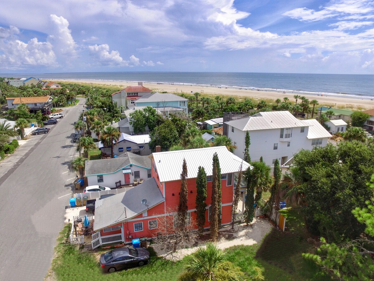 Set in a vibrant coastal community, this image captures the colorful Coral House surrounded by palm trees and greenery. The beach is visible in the background, while the neighborhood streets showcase a mix of residential properties and easy access to the sandy shores.
