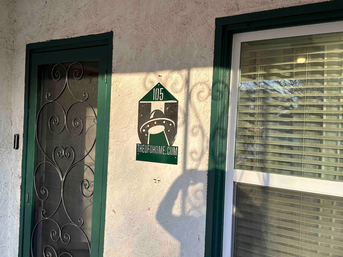 An exterior view of the entrance features a decorative wrought iron screen door with intricate patterns. A house number sign, displaying '105', is mounted on the wall alongside a logo, while sunlight casts shadows on the textured surface.