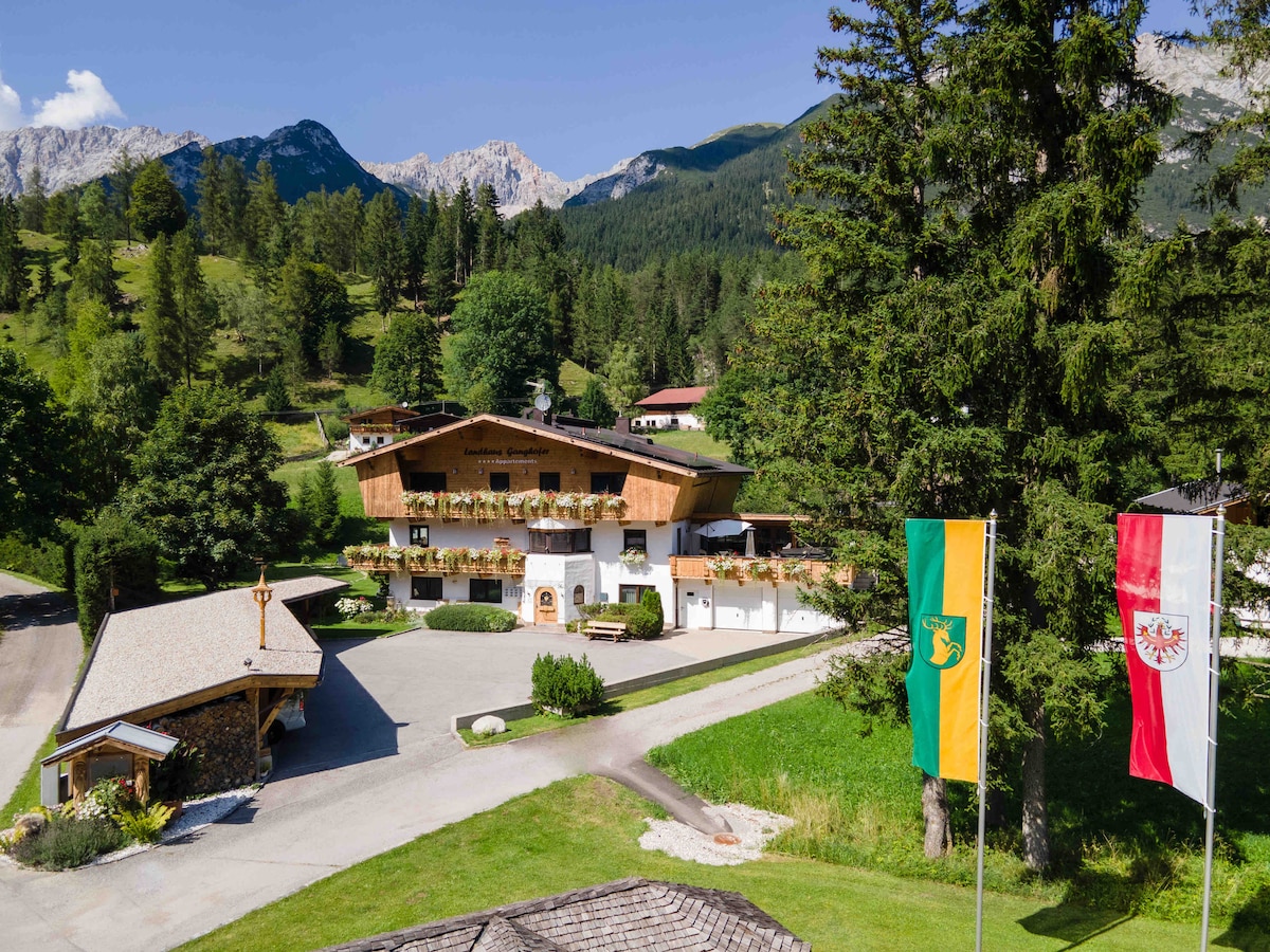 A welcoming alpine residence is set against a backdrop of mountains and lush greenery. The building features multiple balconies adorned with flowers, while two flags flutter gracefully in front. The area is surrounded by trees, enhancing the serene environment.