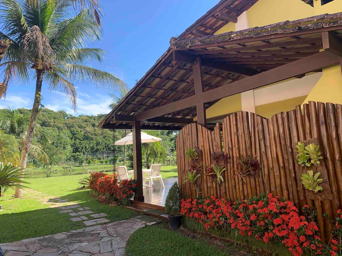A charming porch is framed by a wooden fence adorned with plants, leading to a well-maintained garden. Sun loungers and a parasol are visible on the deck, with vibrant flowers adding color to the lush green surroundings. A serene view of the lake is seen in the background.