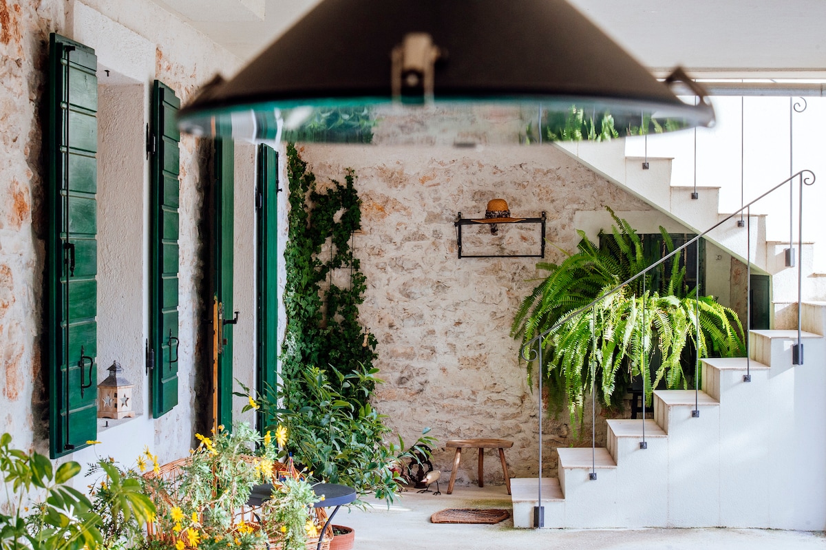 The entrance area displays natural stone walls adorned with green shutters. A lush fern and potted plants enhance the welcoming ambiance. A staircase, bordered by decorative railings, leads to the upper level, while a wooden stool adds a touch of rustic charm.