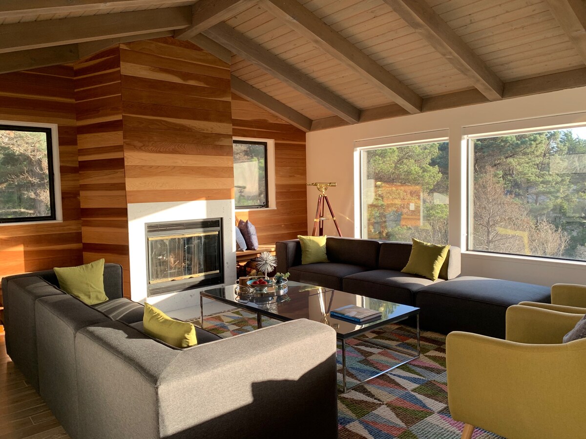 A spacious living area features high vaulted ceilings and large windows that offer views of the surrounding landscape. Comfortable sofas, complemented by accent cushions, are arranged around a glass coffee table. A modern fireplace adds a touch of warmth to the room's inviting design.