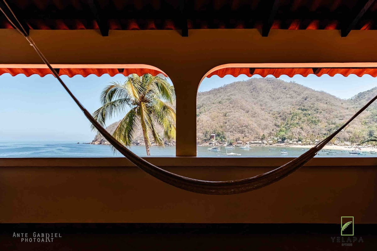 A hammock is suspended between two arched openings, offering a view of Banderas Bay and surrounding mountains. Palm trees are visible in the foreground, framing the coastal landscape and boats in the water.