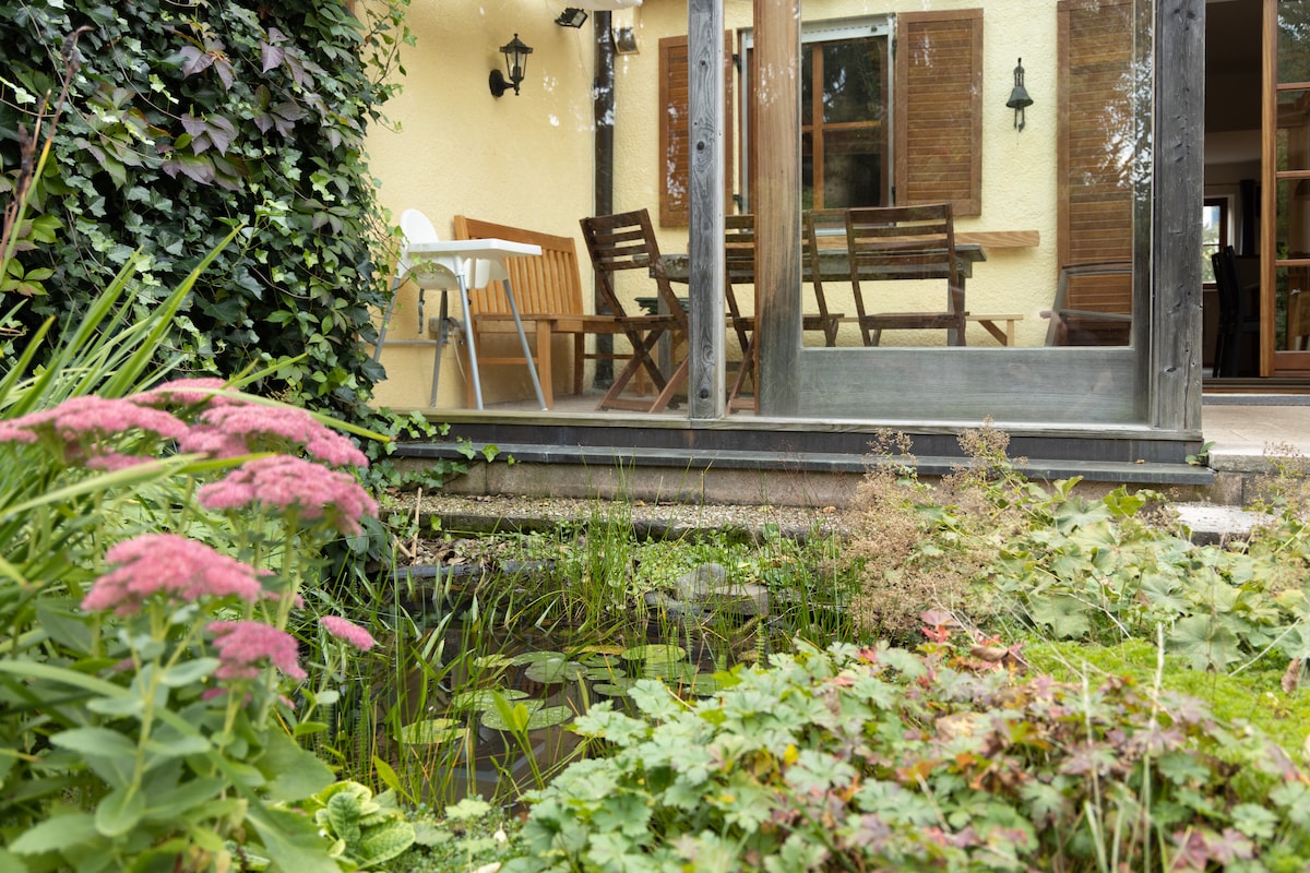 A private terrace is visible through large glass doors, framed by a vibrant garden featuring lush greenery and a small pond. A wooden dining table and chairs are arranged on the terrace, inviting outdoor dining or relaxation.