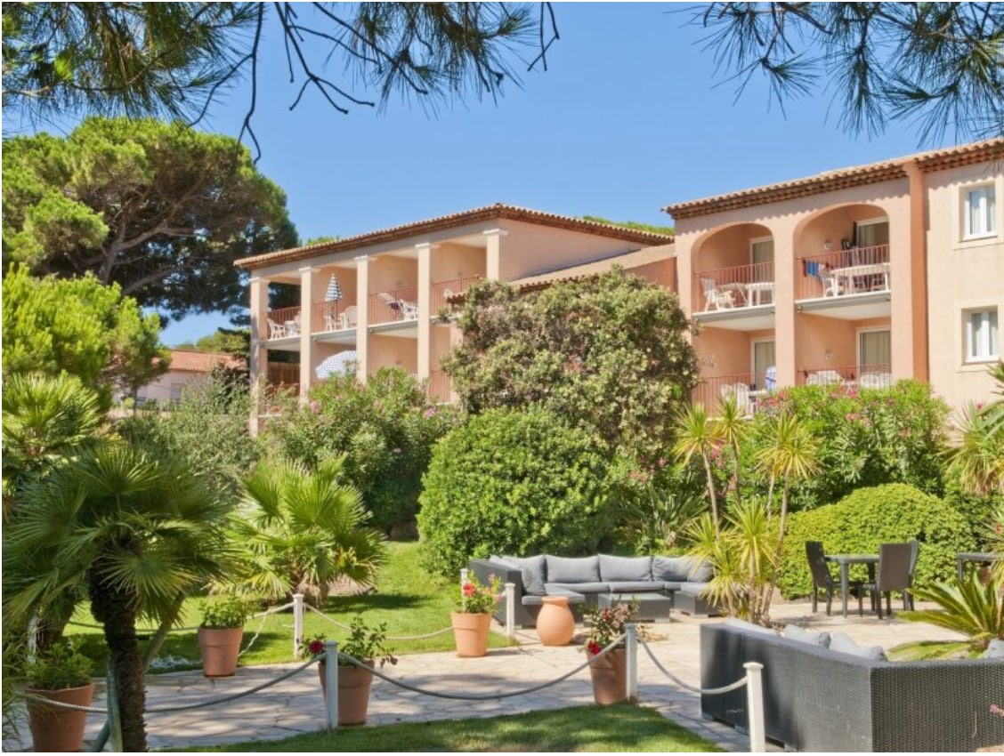 A serene residential building is framed by lush greenery and palm trees. The two-story structure features balconies with railings, while a landscaped garden includes seating areas and potted plants, inviting relaxation in a natural setting.