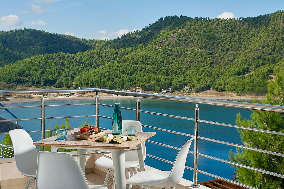 An outdoor dining area is showcased on a balcony, featuring a wooden table surrounded by white chairs. The view reveals a serene landscape of hills covered in lush greenery, reflecting on calm waters. Natural light enhances the inviting ambiance of this private space.