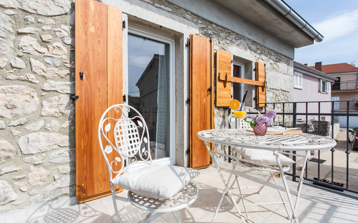 A charming balcony is featured with a small round table and two white wrought iron chairs. The table is adorned with a book and a vase of flowers. Wooden shutters frame the glass door, complementing the natural stone wall behind.