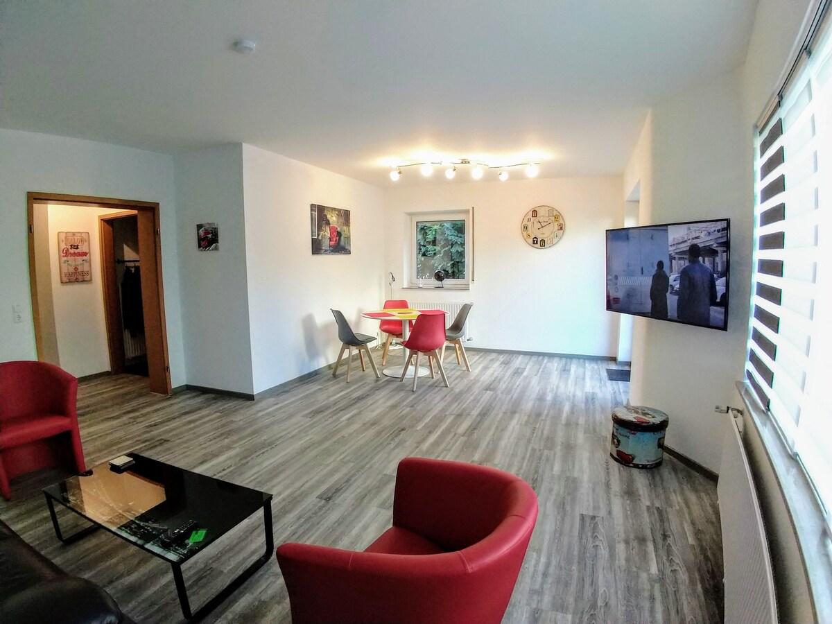 A spacious living area features modern furniture, including a dark coffee table and two red armchairs. A dining table with colorful chairs is positioned near a wall-mounted TV. Soft lighting from above complements the neutral wall and floor design.