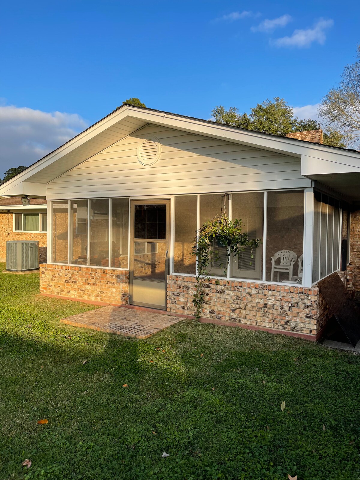A screened-in patio is featured, surrounded by brick walls and a neatly maintained lawn. Lightly shaded by trees, it offers a view of the neighborhood. Two white chairs are visible inside the patio, inviting relaxation in a serene environment.