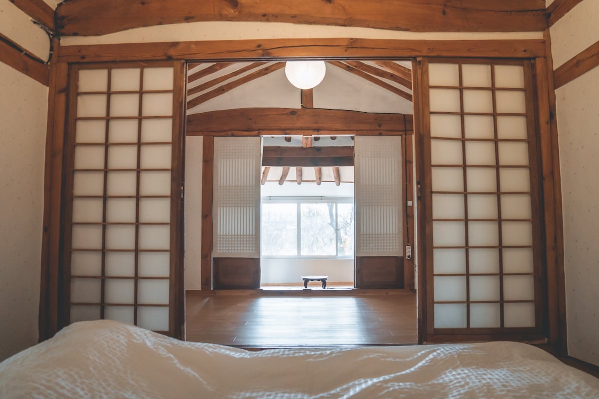 A spacious room features traditional sliding doors with wooden frames, allowing natural light to illuminate the interior. The room's high wooden ceiling creates an airy atmosphere, while a simple low table can be seen in the distance, framed by a large window showcasing the outside view.