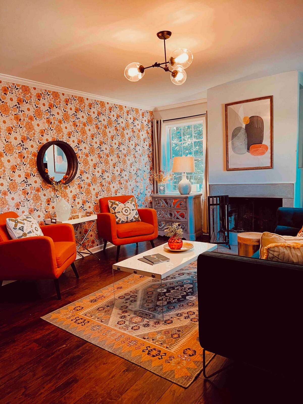 A comfortable living room is highlighted by two red armchairs and a black sofa, arranged around a coffee table. A colorful area rug lies beneath. The wall is adorned with floral wallpaper, and a window allows natural light to fill the space.