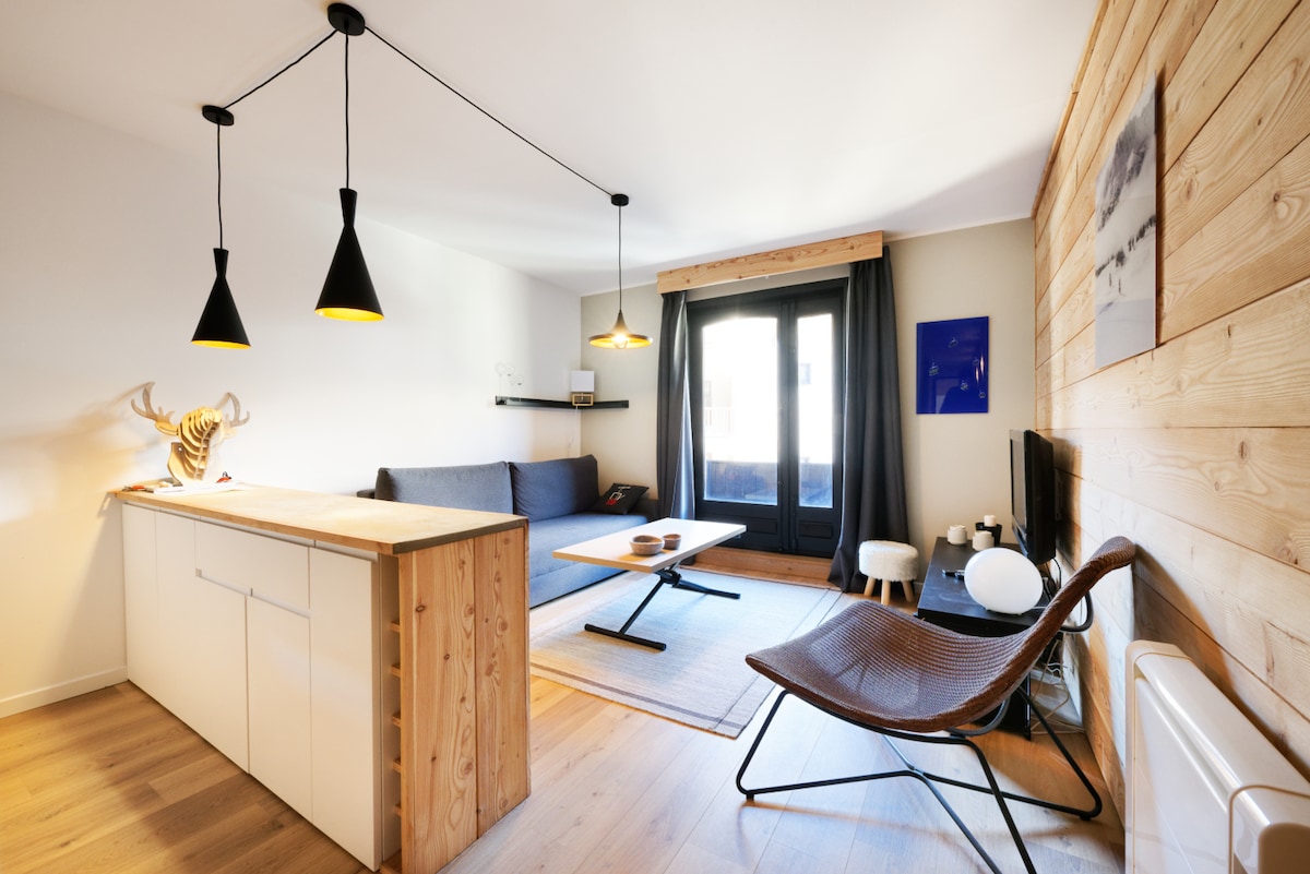 A modern living area features a cozy sofa and a wooden coffee table, complemented by a sleek TV unit. Two pendant lights hang above the space, and large windows allow natural light to fill the room. Rustic wood paneling adds character to the walls.