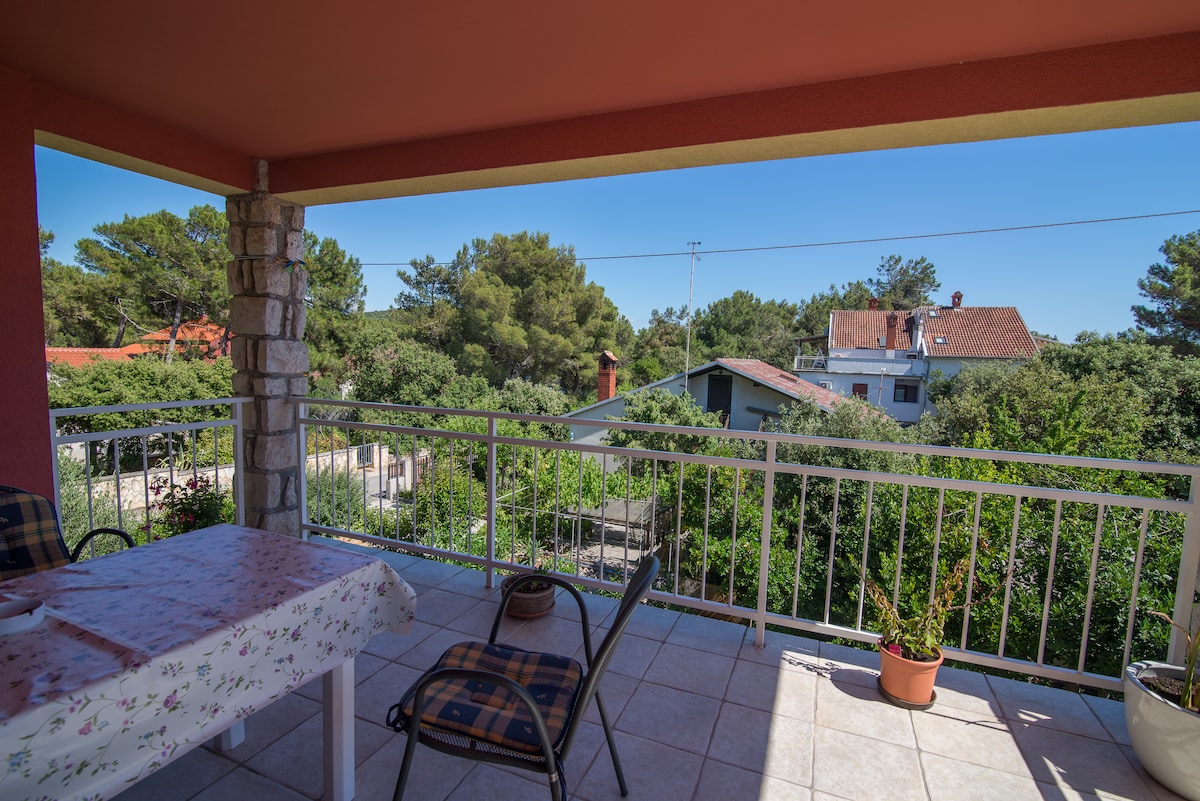 A covered balcony is presented with a small table and two chairs, surrounded by lush greenery. The view includes trees and nearby buildings, enhancing the setting's natural charm. The tiled floor is brightened by sunlight, creating a welcoming space for relaxation.