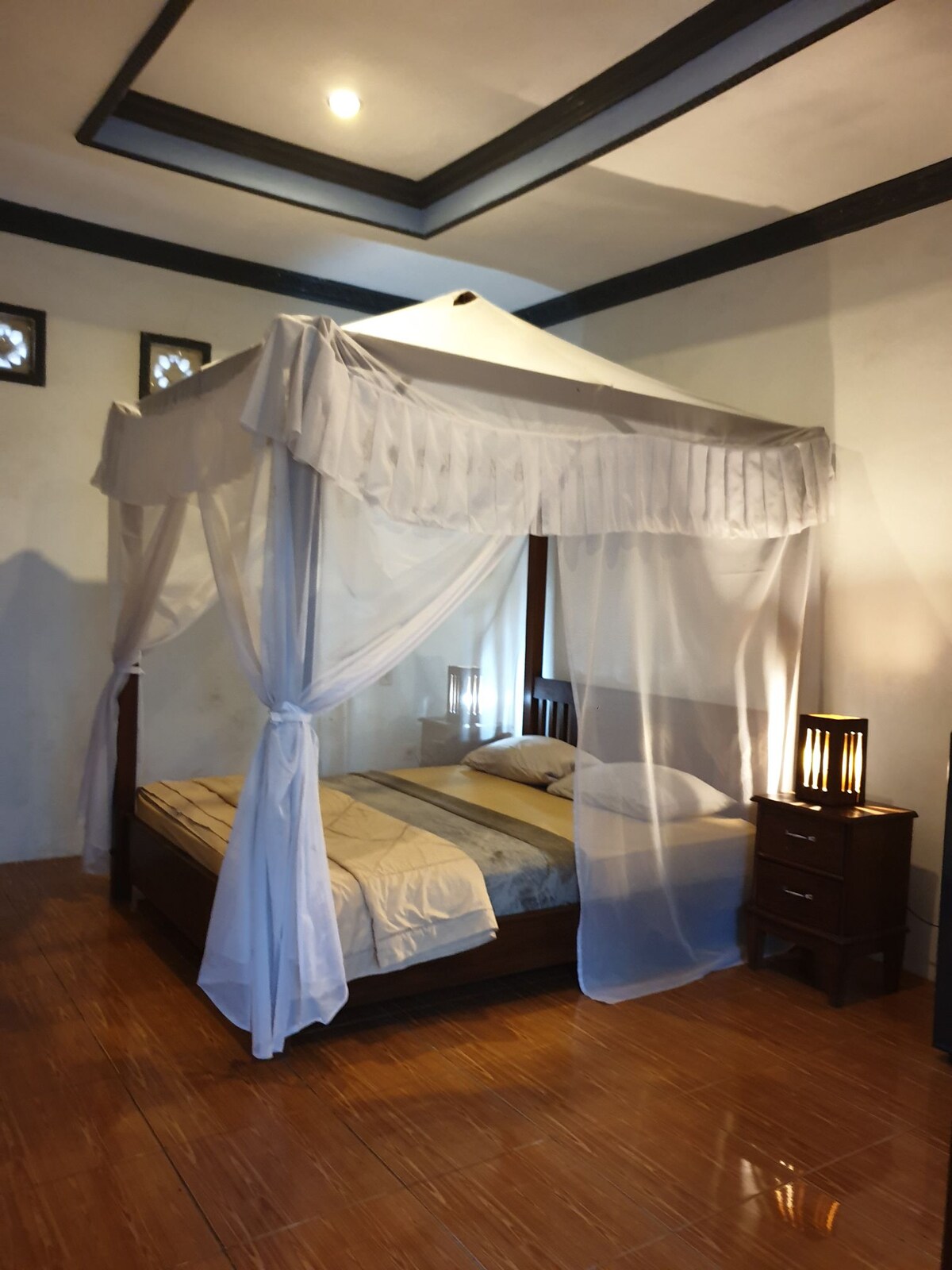 A spacious bed is covered with a light-colored linen and draped with a sheer canopy. Soft lighting from bedside lamps creates a gentle glow, while wooden flooring adds warmth to the room, which features simple decorative elements and neutral wall tones.