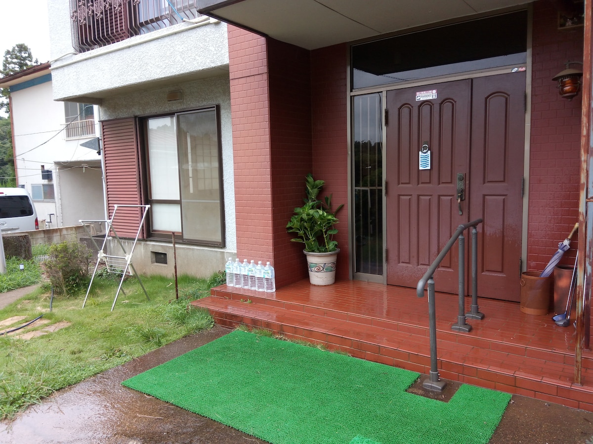 A welcoming entrance features a set of double doors framed by a potted plant and a small seating area. The entrance is complemented by a green mat flooring and a neatly arranged set of water bottles by the steps, all set against a rustic brick facade.