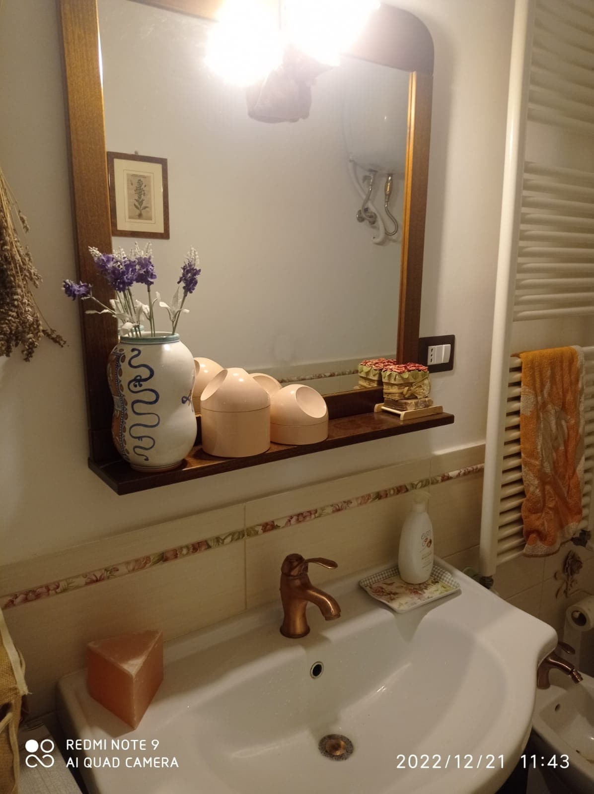 A bathroom sink area features a sleek white basin and a decorative wooden shelf above it. On the shelf, a ceramic vase with lavender, two soft-colored containers, and a small wooden box are displayed. Warm lighting enhances the inviting ambiance.