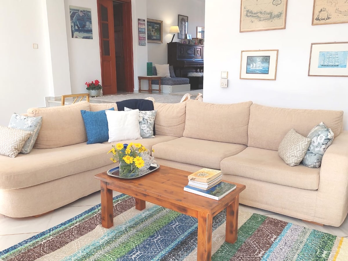 A spacious living area is highlighted by a large, light-colored sectional sofa adorned with decorative pillows in various patterns. A wooden coffee table holds a floral arrangement and books, while a colorful rug adds texture to the floor.