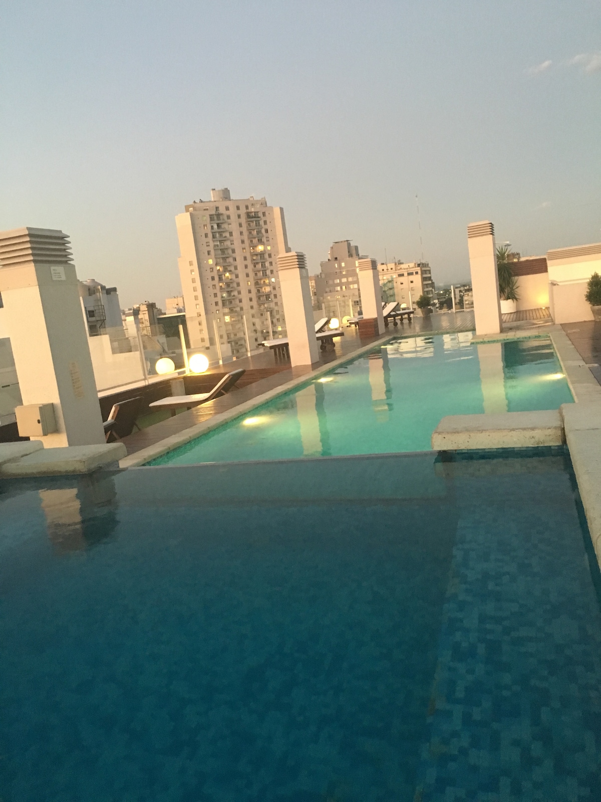 An outdoor pool is observed on a rooftop, surrounded by modern buildings. The clear water reflects the sky, while lounge chairs and illuminated spheres are positioned along the edge. A serene atmosphere is enhanced by the open space and urban view.