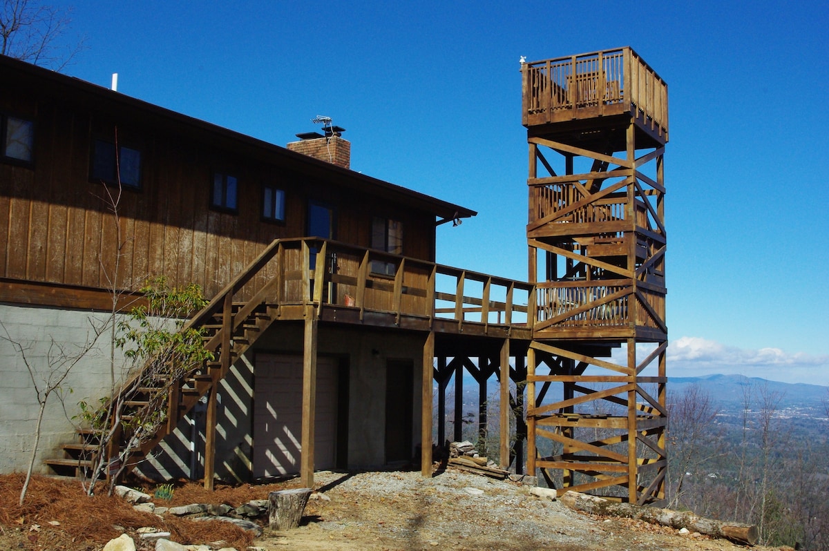 A wooden observation tower stands adjacent to the cabin, offering elevated views of the surrounding landscape. A staircase leads to the tower's entrance, while a wide deck wraps around the cabin, providing access to the main entrance and enhancing outdoor enjoyment.