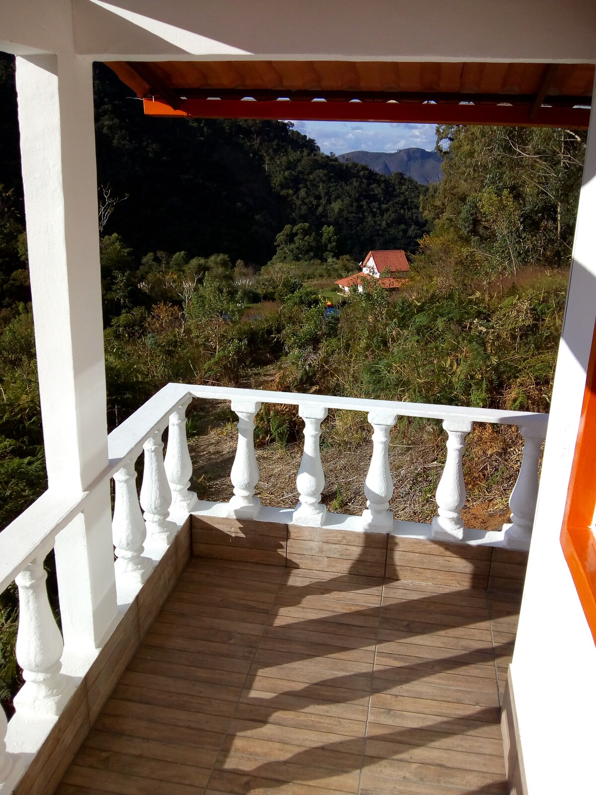 A balcony features white railing and terracotta tiled flooring, overlooking a natural landscape. Lush greenery and distant mountain peaks are visible, along with a glimpse of a red-roofed house nestled in the valley below under a clear sky.