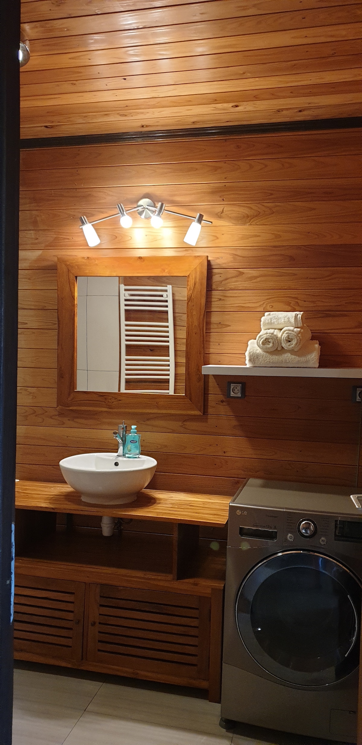 A compact bathroom featuring warm wooden paneling and a light fixture above a sink. The sink is set on a wooden countertop with open storage below. A modern washing machine occupies the corner, and a decorative sculpture is placed on a shelf above the sink.