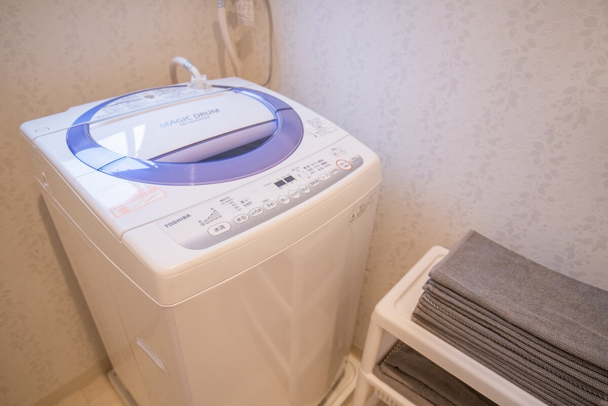 A compact washing machine is positioned next to a stack of neatly folded gray towels on a white shelf. The machine features a blue control panel and is situated against a softly patterned wall, enhancing the functionality of the laundry area.