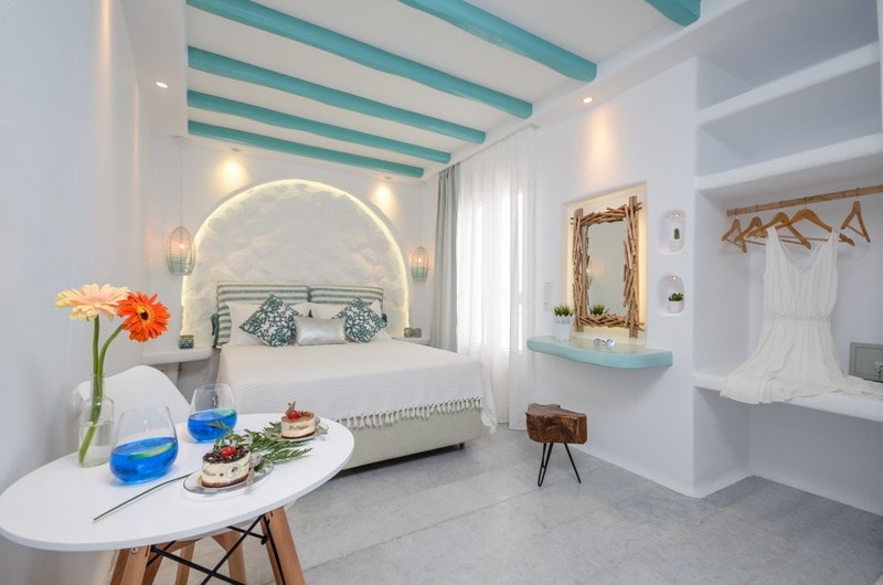 The room features a spacious bed with a decorative headboard and colorful pillows. Natural light filters through sheer curtains, while a white table with drinks and snacks is positioned near the bed. A mirror and hanging clothes add practicality, complemented by a floral touch.