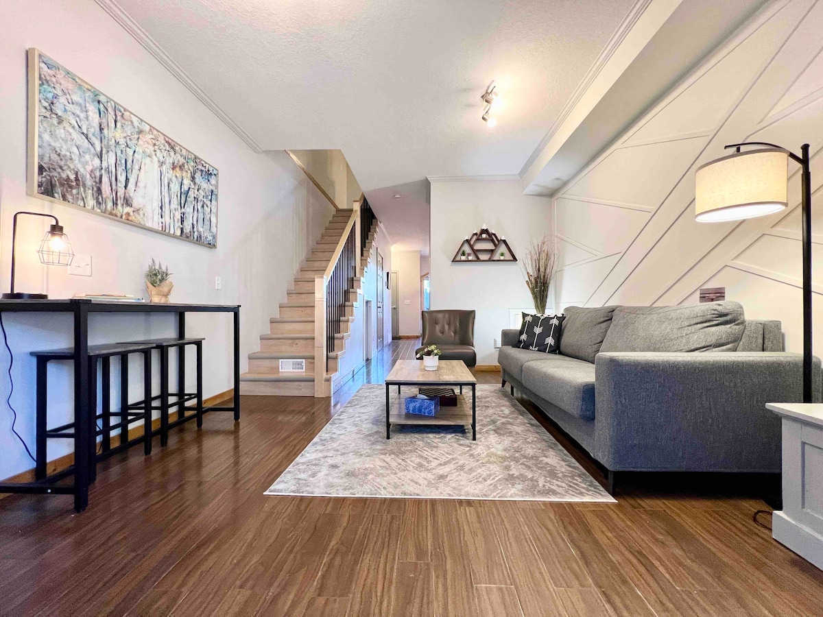 The living area showcases a spacious gray sectional sofa positioned on a soft area rug. A wooden coffee table sits centrally, complemented by a sleek side table with a lamp. A dining-like setup with bar stools and artwork adds a touch of character to the space.