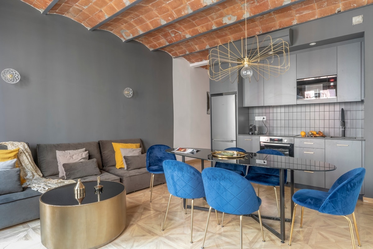 An open living area showcases a modern kitchen and dining space. A grey sofa with colorful cushions complements a round coffee table. Four blue velvet chairs surround a sleek dining table, and a stylish light fixture hangs from the ceiling, accentuating the brick archways.
