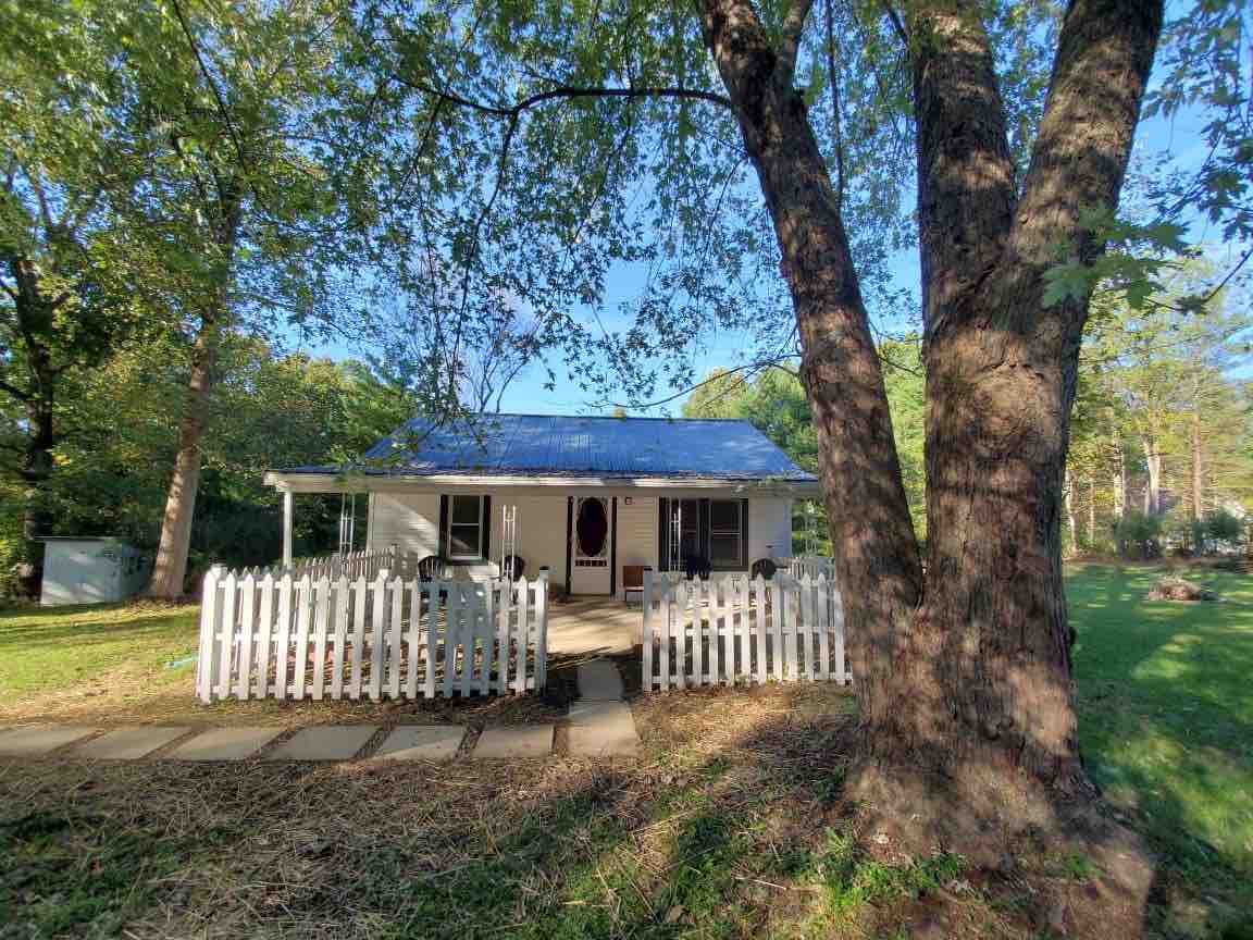 A quaint home is framed by mature trees and a white picket fence. The front porch features several chairs, inviting relaxation. Sunlight filters through the leaves, highlighting the natural surroundings and the pathway leading up to the entrance.