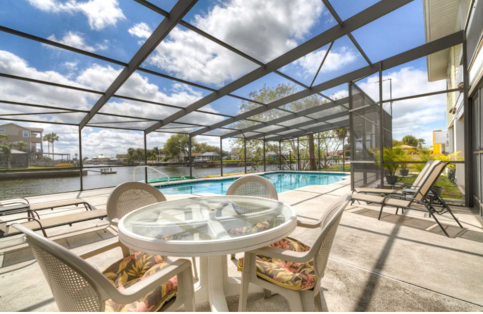 An outdoor patio area features a round table surrounded by four chairs, set against a backdrop of a private pool and waterway. A spacious pool deck is visible, with two lounge chairs positioned for relaxation, all under a screened enclosure that allows for unobstructed views.
