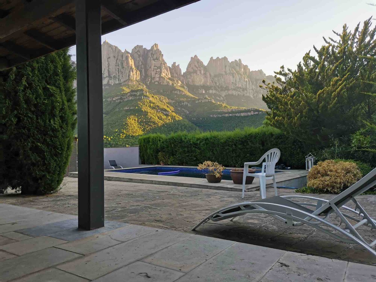 A tranquil outdoor area showcases a private pool, surrounded by lush hedges and mountains in the background. A sun lounger and a small vase of flowers add to the serene atmosphere, while natural stone flooring complements the setting.