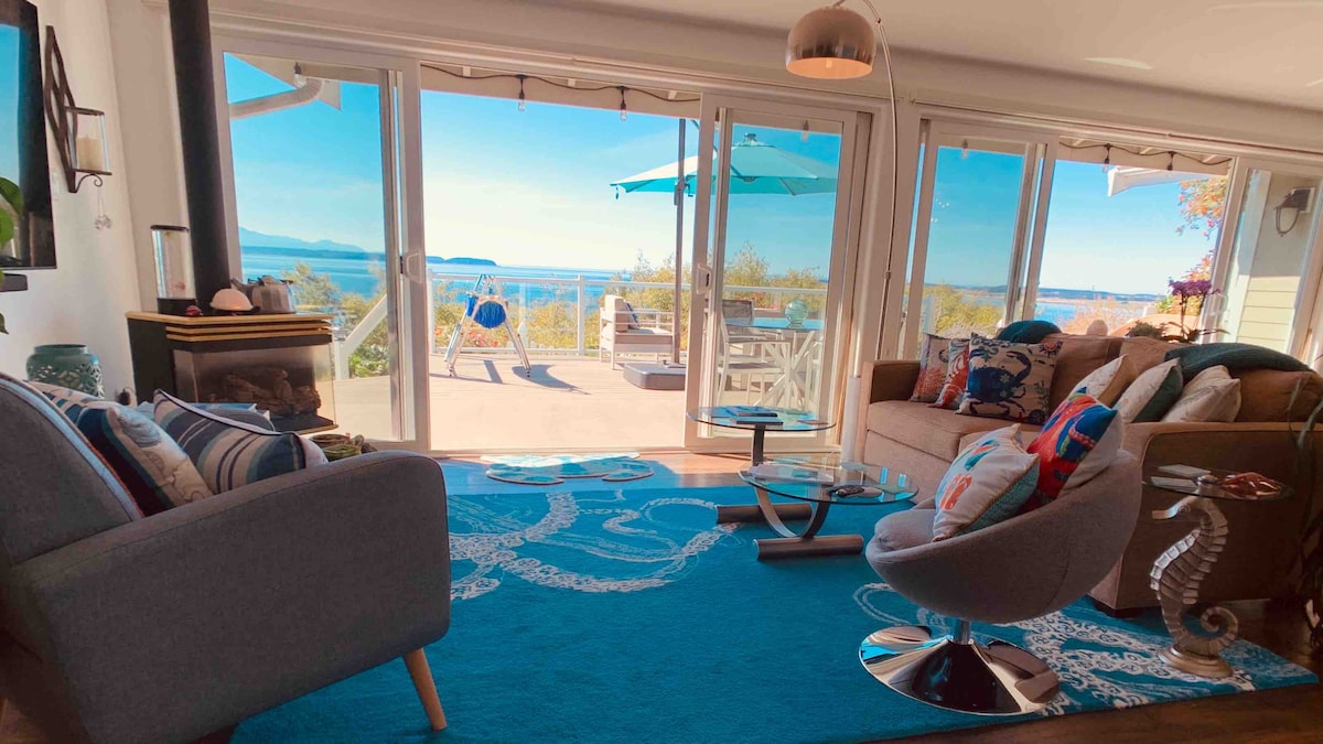 A sunlit living area is showcased, featuring comfortable seating arrangements on a vibrant blue area rug. Large glass doors offer an unobstructed view of the outdoor deck overlooking Admiralty Inlet and the surrounding landscape.