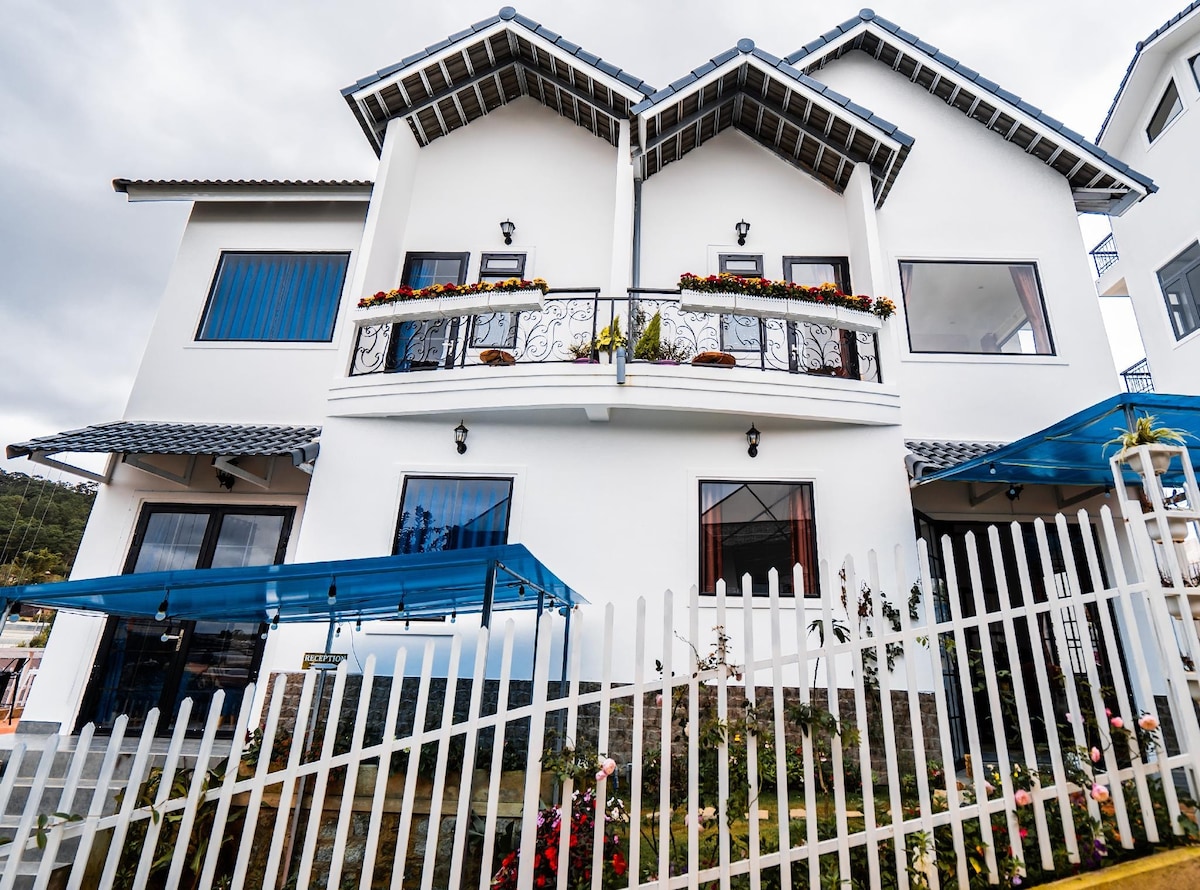A two-story white house features a distinctive gabled roof and blue accents. Balconies are adorned with potted plants, and large windows offer views of the surroundings. A white picket fence surrounds the property, highlighting the vibrant flowers in the garden.