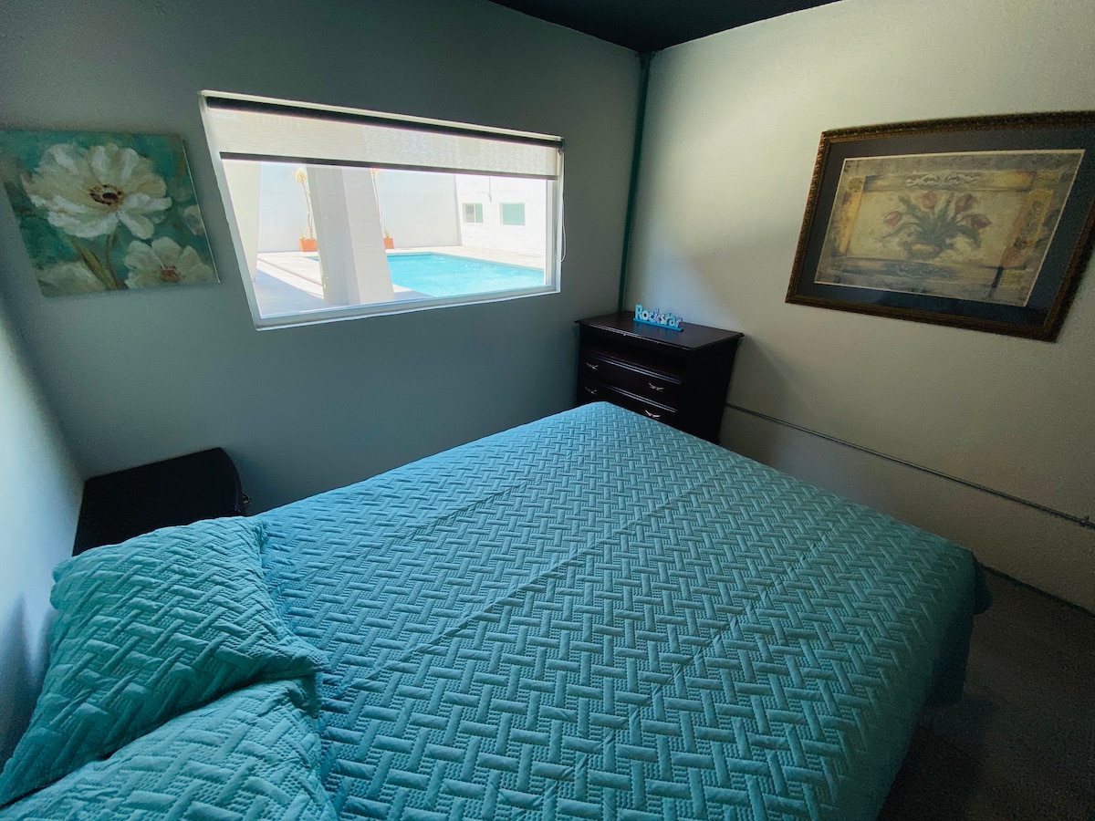 A cozy bedroom features a spacious bed covered with a textured aqua blanket. Natural light enters through a large window, revealing a glimpse of a pool outside. A dark wooden dresser is positioned against the wall, and a wall art piece adds a touch of decor.