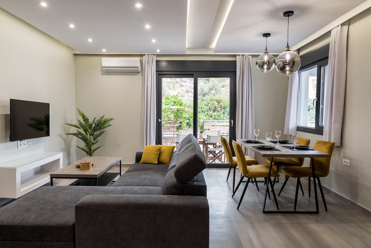 A modern living area is depicted, featuring a spacious gray sofa positioned beside a sleek coffee table. A dining table with stylish chairs accommodates dining needs. Natural light enters through large glass doors that open to a terrace, complemented by green foliage outside.