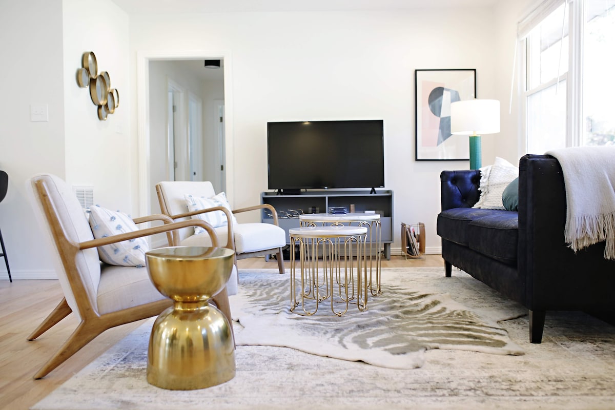 The living area features a contemporary design with a dark blue sofa and light-colored armchairs. A round brass table and a sculptural coffee table are positioned on a soft area rug. A flat-screen TV is mounted on a light gray console under a bright lamp.