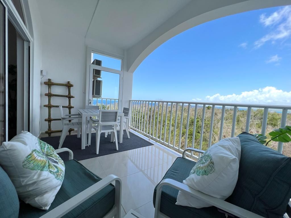 A private terrace features outdoor seating with cushions for comfort, along with a dining table for four. The view extends from the terrace, showcasing the surrounding greenery under a bright blue sky.