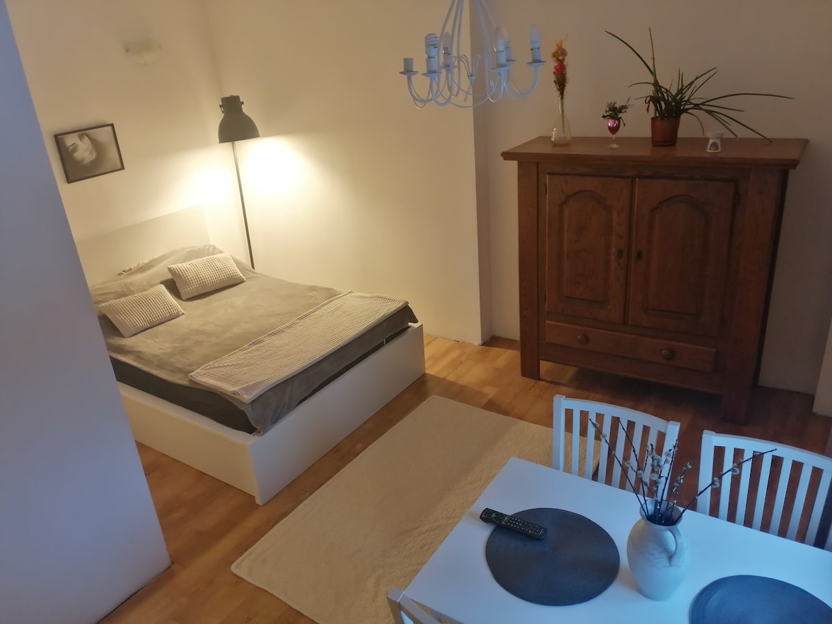 A cozy sleeping area is presented, featuring a spacious bed with soft linens. A wooden cabinet stands against the wall, and a small dining table with white chairs is set nearby. Warm light from a chandelier creates a comfortable ambiance.